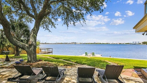 A patio with lounge chairs overlooking a body of water, a dock, and a grassy area. - Amazing Bay by Ryson Vacation Rentals (Galveston)