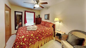 A bedroom with a bed, a chair, a lamp, a ceiling fan, a window with curtains, and a door. - Pinon Park C11 by Ruidoso Vacation Rentals (Ruidoso)