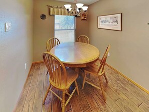 A dining area with a round wooden table and six chairs, a window with blinds, a chandelier, and a framed picture on the wall. - Pinon Park C11 by Ruidoso Vacation Rentals (Ruidoso)