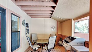 A covered patio with a round table and four chairs, a grill, and a window with blinds. - Pinon Park C11 by Ruidoso Vacation Rentals (Ruidoso)