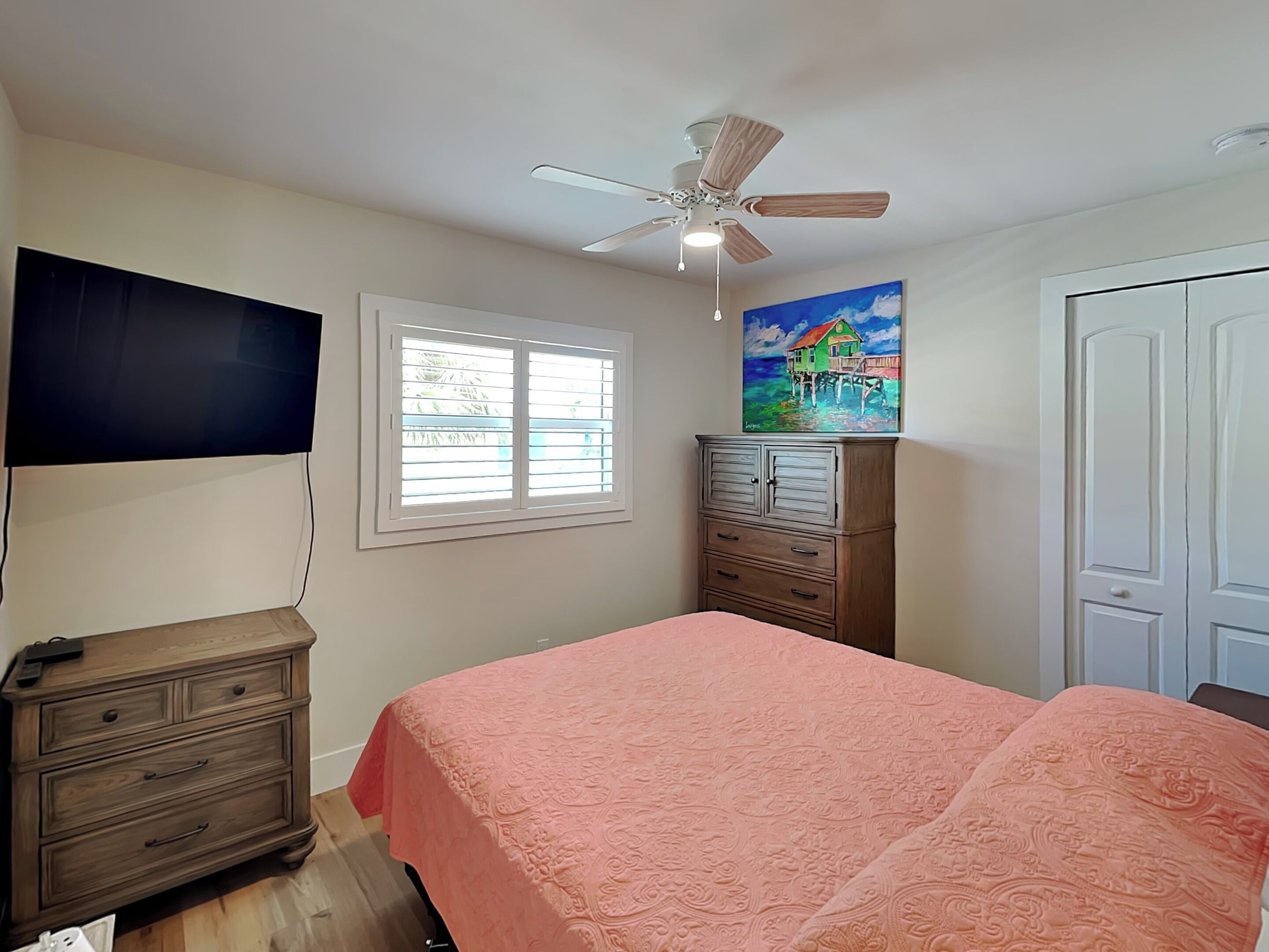 Une chambre à coucher avec un lit, un ventilateur de plafond, une fenêtre, une commode et un tableau.