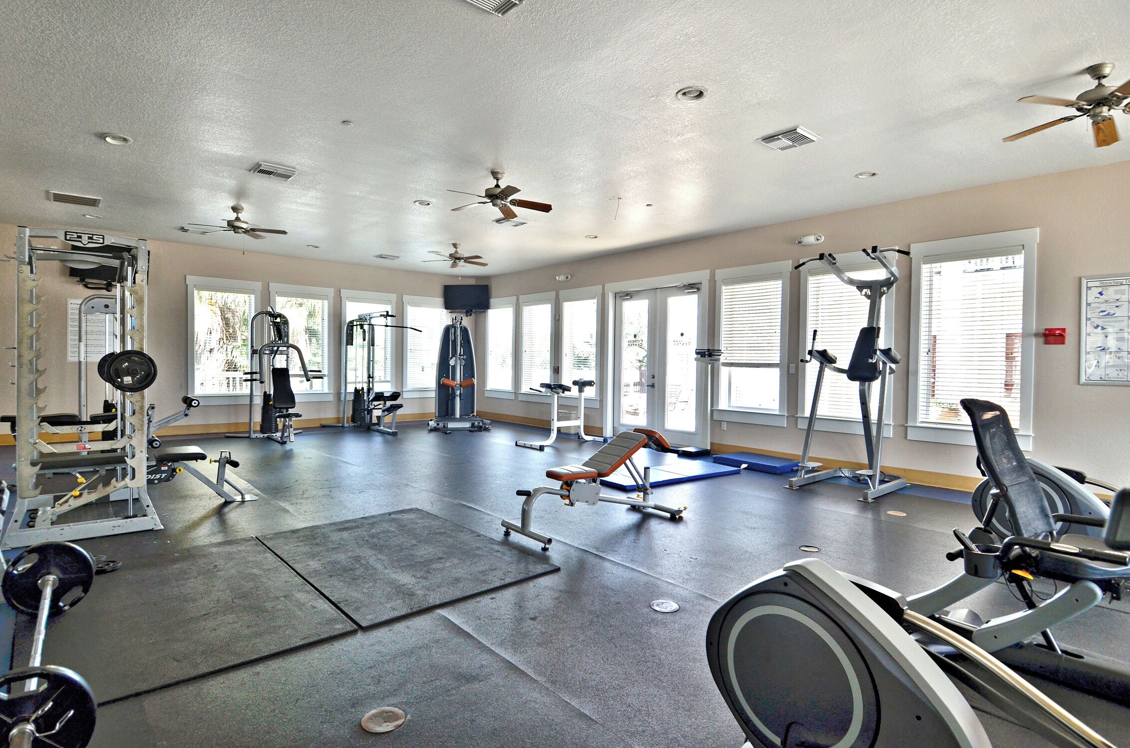 Une salle de sport spacieuse, dotée de divers appareils d’exercice, de grandes fenêtres et de ventilateurs au plafond.