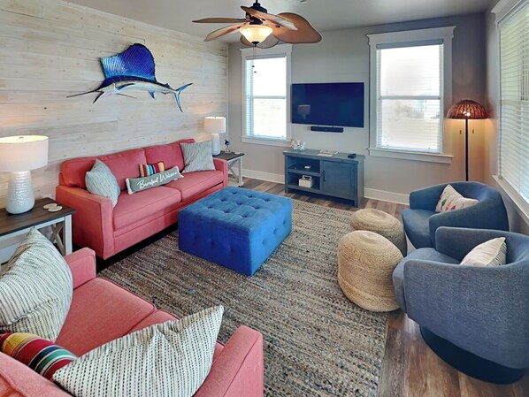 A living room with a red sofa, blue ottoman, and blue armchairs. There is a ceiling fan, a TV, and a decorative fish on the wall. - Beachside Blue Sailfish by Ryson Vacation Rentals (Galveston)