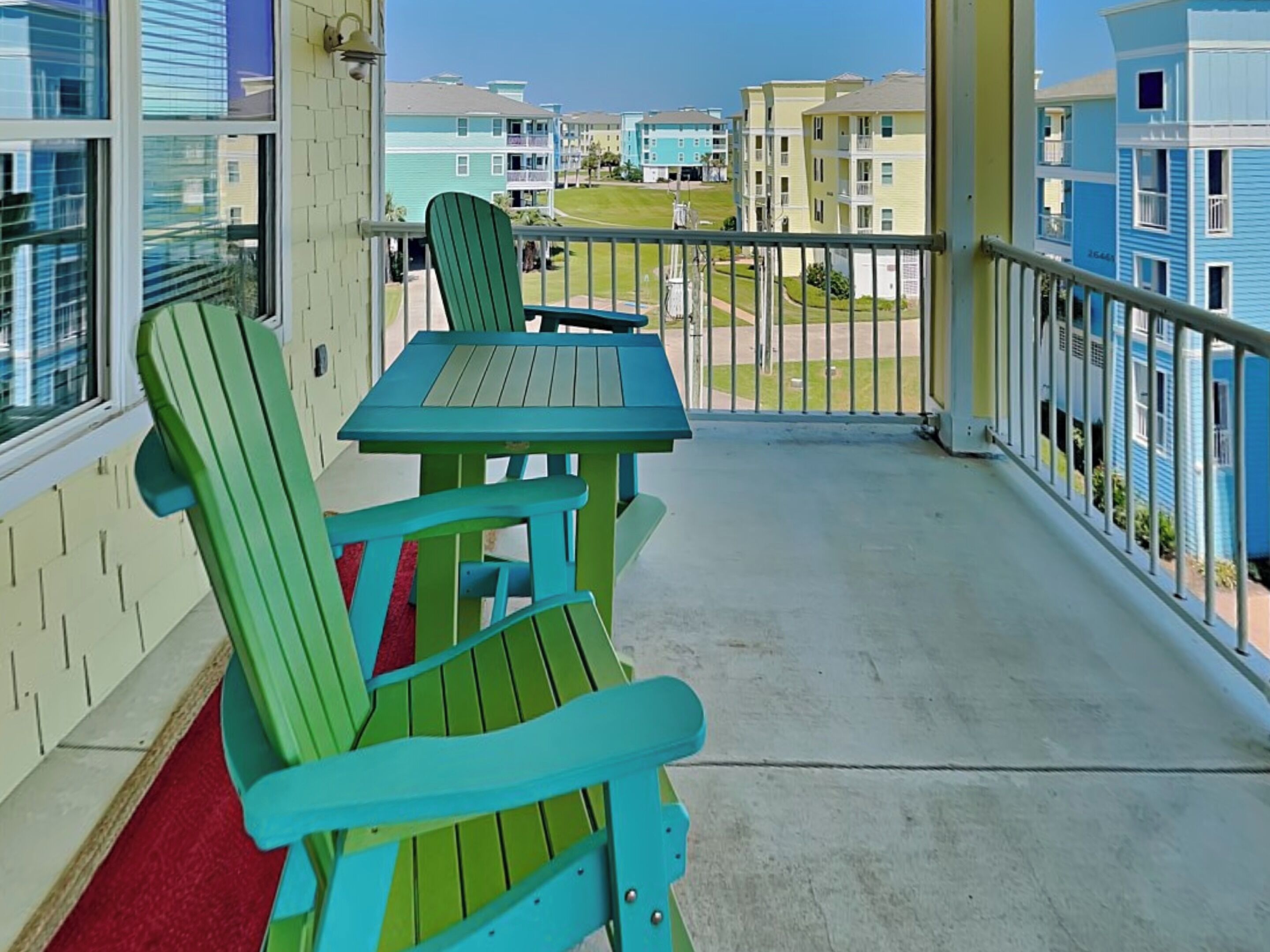 A balcony with a green Adirondack chair and a small table, overlooking a row of colorful buildings.