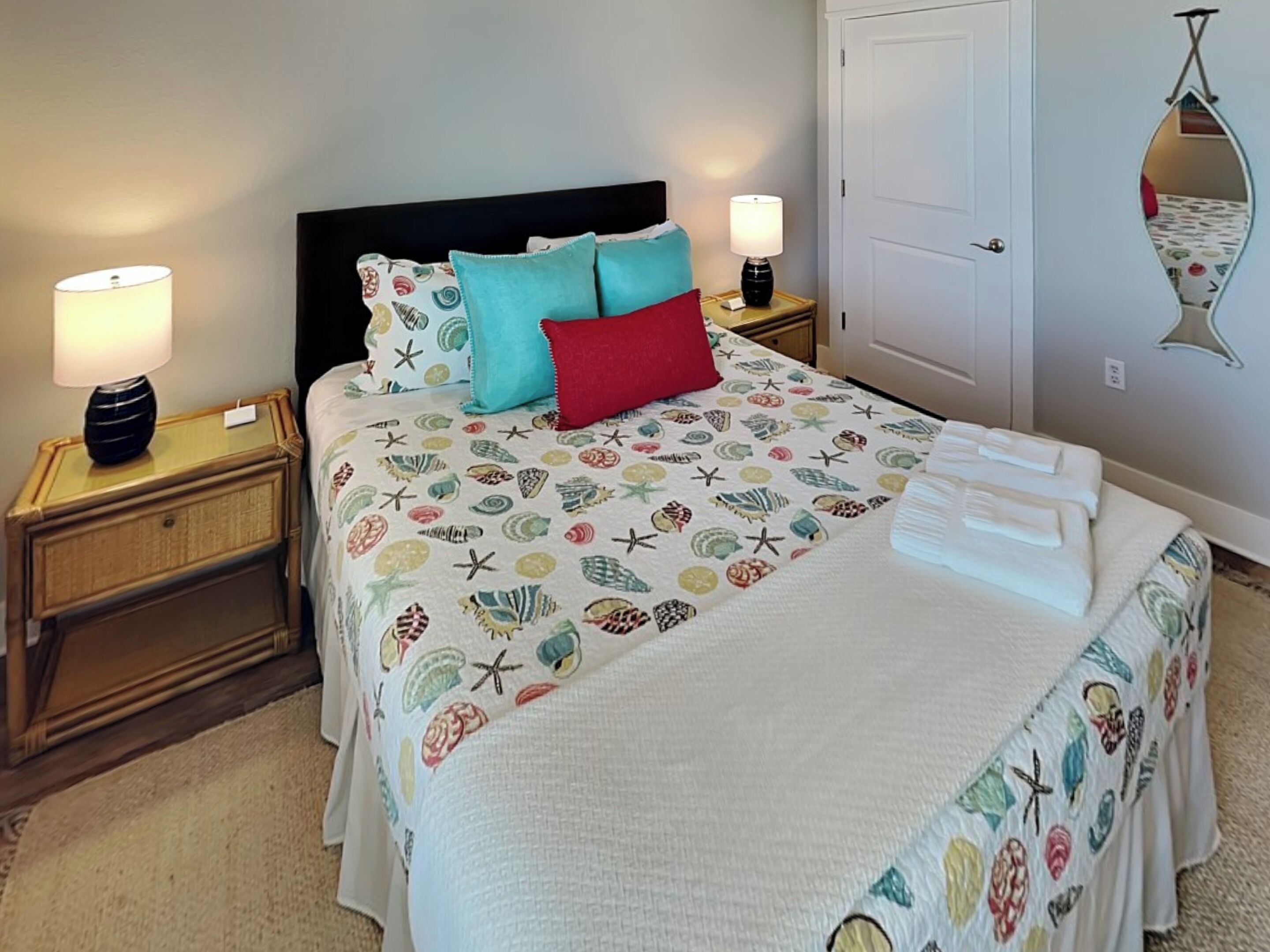 Una cama bien hecha con una colcha estampada, dos almohadas y una manta blanca. Hay dos mesitas de noche con lámparas y un pequeño espejo en la pared.