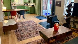 Un salon avec une table basse en bois, un tapis et un vase rouge. Au fond, on aperçoit une cuisine avec des meubles verts et un fourneau.