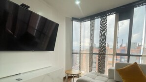 Ein modernes Wohnzimmer mit einem großen Flachbildfernseher an der Wand, einem weißen Sofa mit einem senfgelben Kissen, einem kleinen runden Couchtisch und Blick auf die Stadt durch große Fenster.