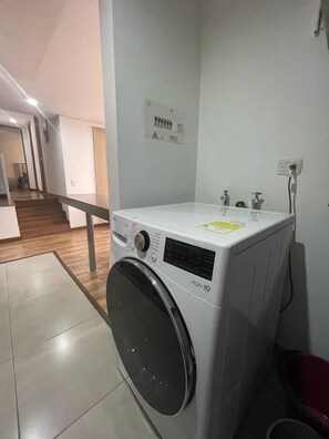 Une petite buanderie moderne, équipée d’un lave-linge à chargement frontal, d’un petit évier et d’une prise de courant au mur.