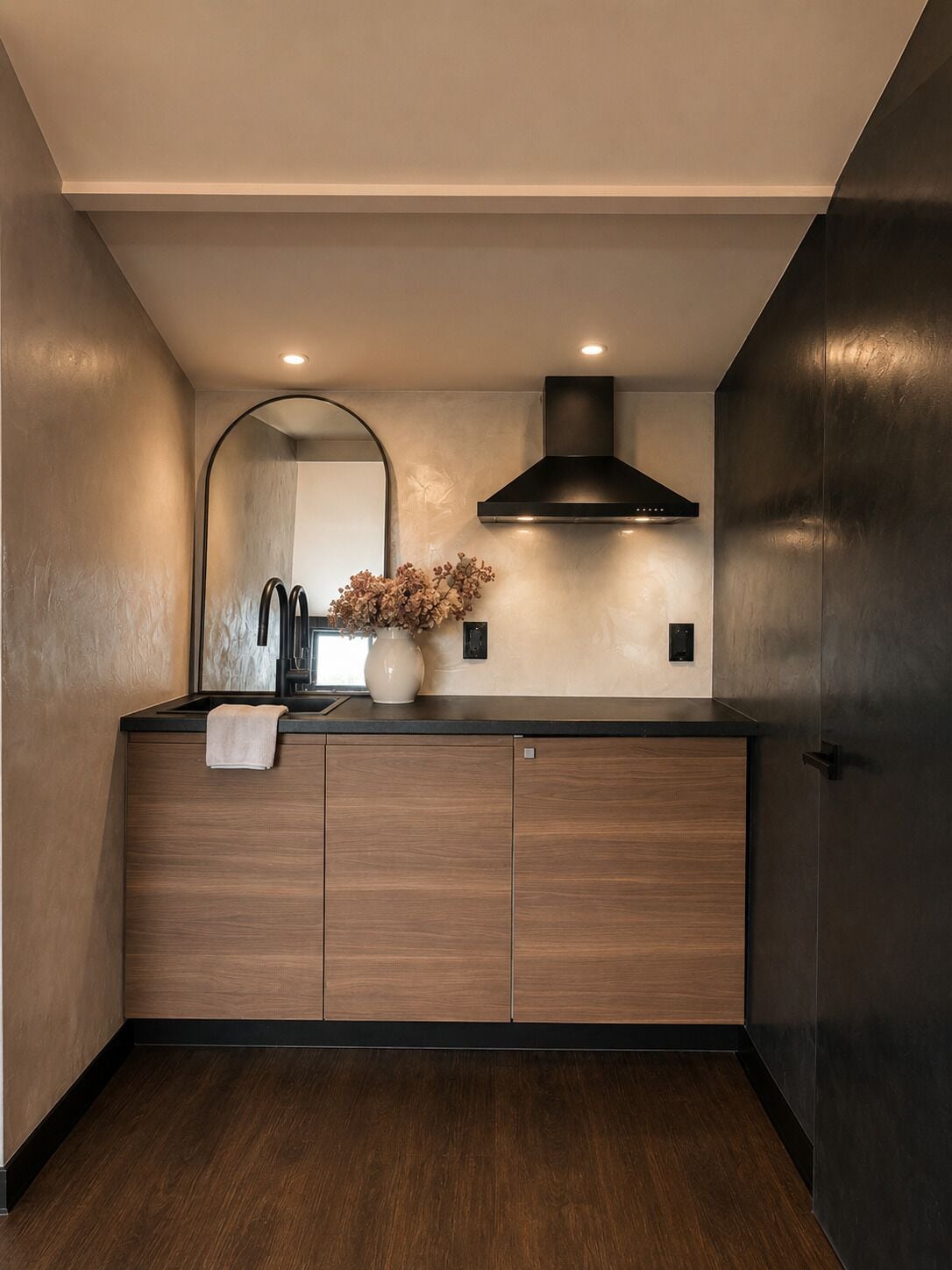 A modern kitchen with wooden cabinets, a black range hood, a mirror, and a vase with dried flowers.