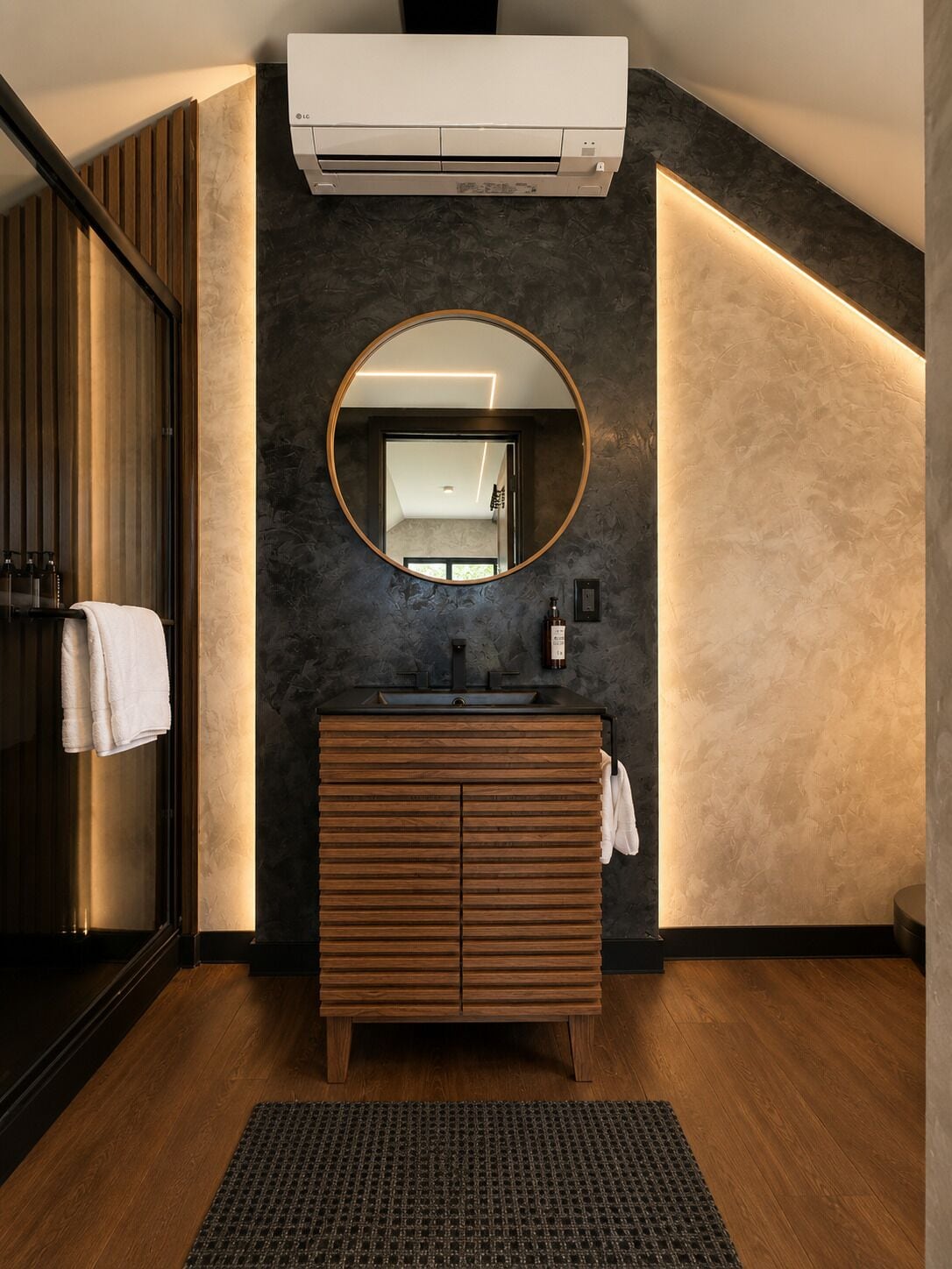 Un baño moderno con tocador de madera, un espejo redondo y un equipo de aire acondicionado integrado.