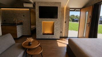 A modern hotel room with a sofa, a small table with a vase, a fireplace, a TV, and a view of the outdoors.