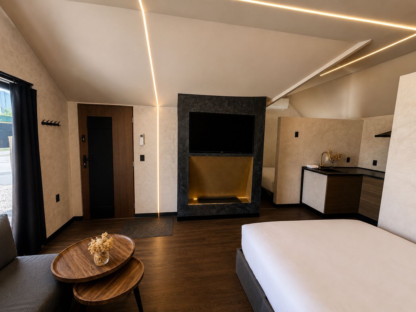Une chambre d’hôtel moderne comprenant un lit, un coin salon avec une table en bois, une cheminée intégrée, une télévision et une kitchenette.