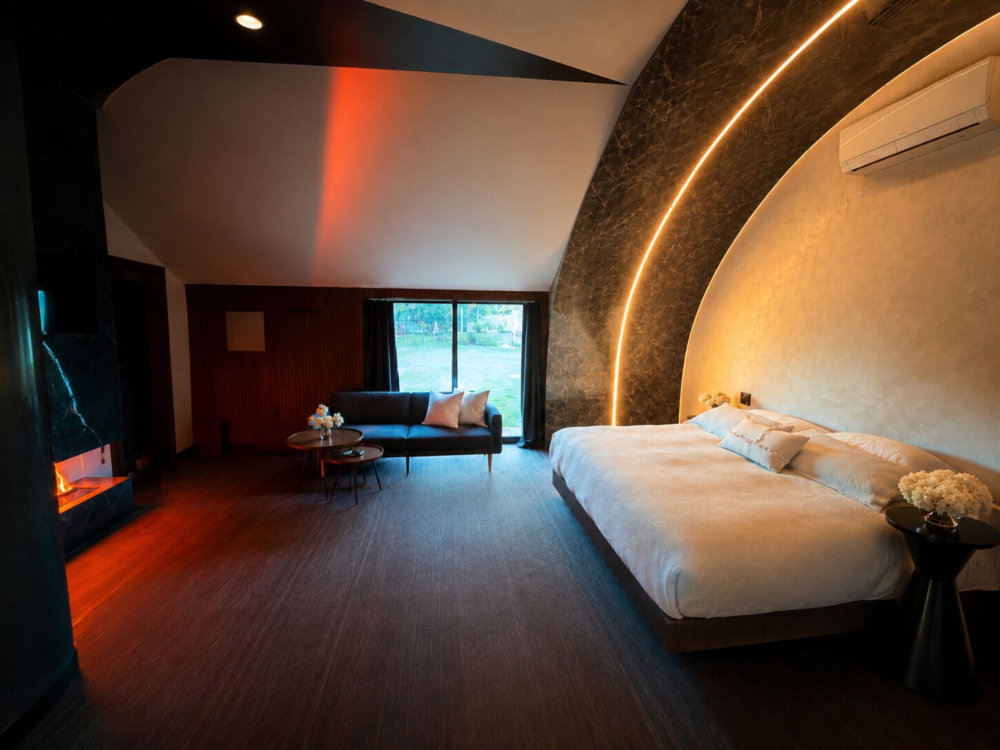 A modern hotel room with a curved ceiling, a large bed, a sofa, a small table, and a view of the outdoors.