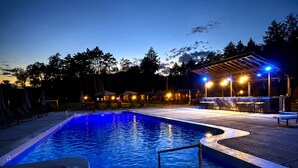 Ein Schwimmbecken im Abenddämmerung mit Liegestühlen und einem überdachten Bereich mit Beleuchtung.