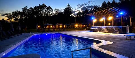 A swimming pool at dusk with surrounding lounge chairs and a covered area with lights.