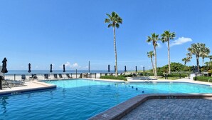 Uma piscina com água azul cristalina, rodeada por palmeiras e espreguiçadeiras, com vista para o oceano.
