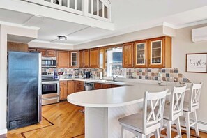 A modern kitchen with wooden cabinets, a stainless steel refrigerator, a large island with bar stools, and a tiled backsplash. - Lakeside Retreat on Saratoga Lake (Saratoga Springs)