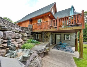 A two-story wooden cabin with a stone foundation, a balcony, and a patio area with outdoor furniture. - Modern Retreat | Near Hunter & Windham | Dog-Friendly (Jewett)