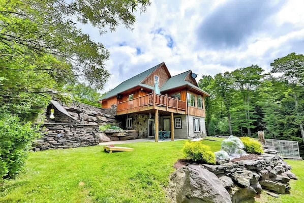 A two-story log cabin with a stone foundation, a wooden deck, and a stone retaining wall. - Modern Retreat | Near Hunter & Windham | Dog-Friendly (Jewett)