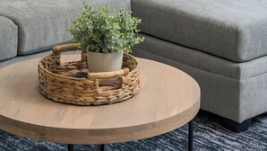 Ein modernes Wohnzimmer mit einem grauen Sofa, bunten Kissen, einem gewebten Couchtisch und einer Wandleuchte.