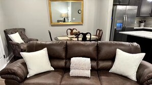 Un sofá de cuero marrón con cojines blancos, una mesita con papeles, una silla y una zona de cocina con un refrigerador.