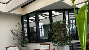Een overdekt terras met rieten stoelen en grote potplanten.