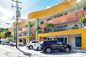 A multi-story hotel with a yellow facade, green pillars, and balconies with plants. There are several cars parked in front.