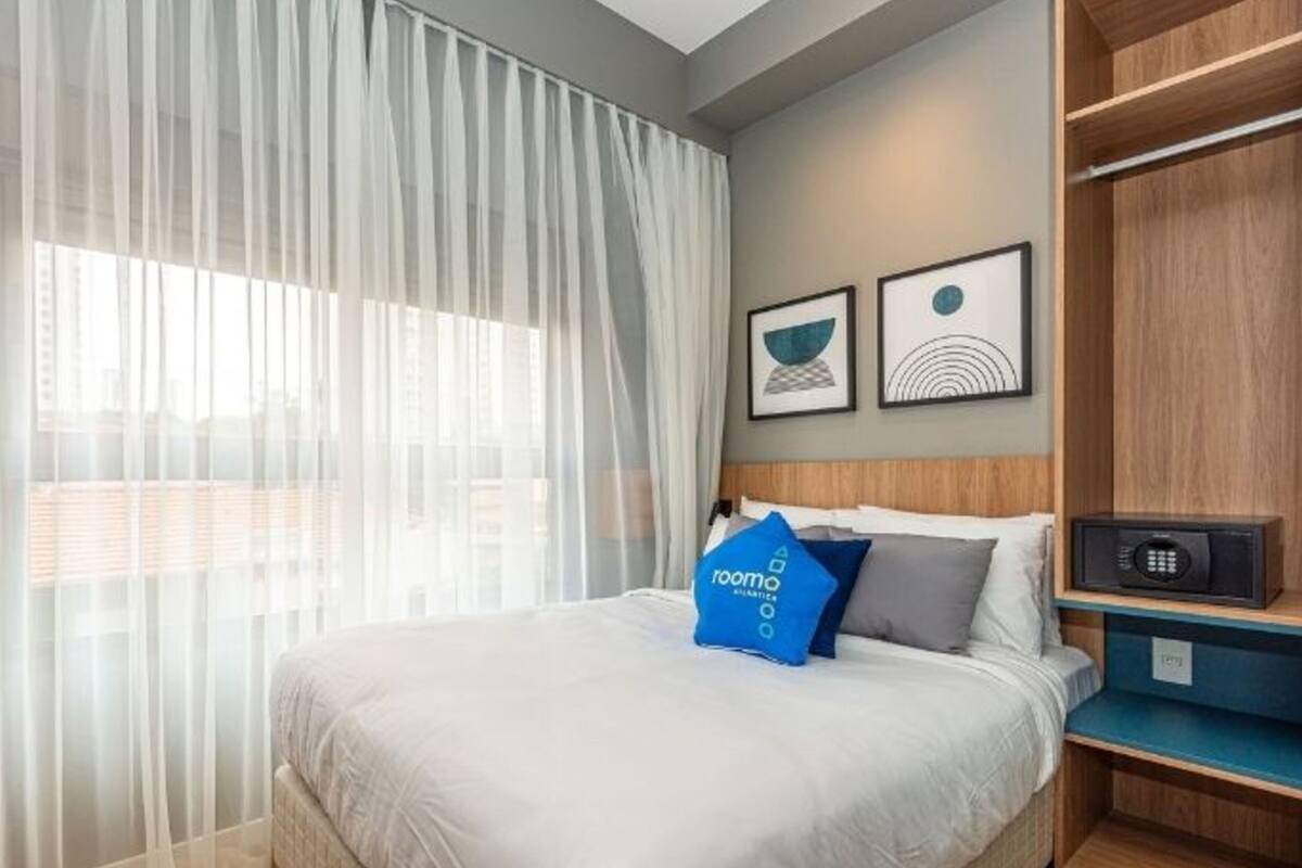 A hotel room with a bed, a window with sheer curtains, a wall-mounted shelf with a speaker, and two framed artworks.