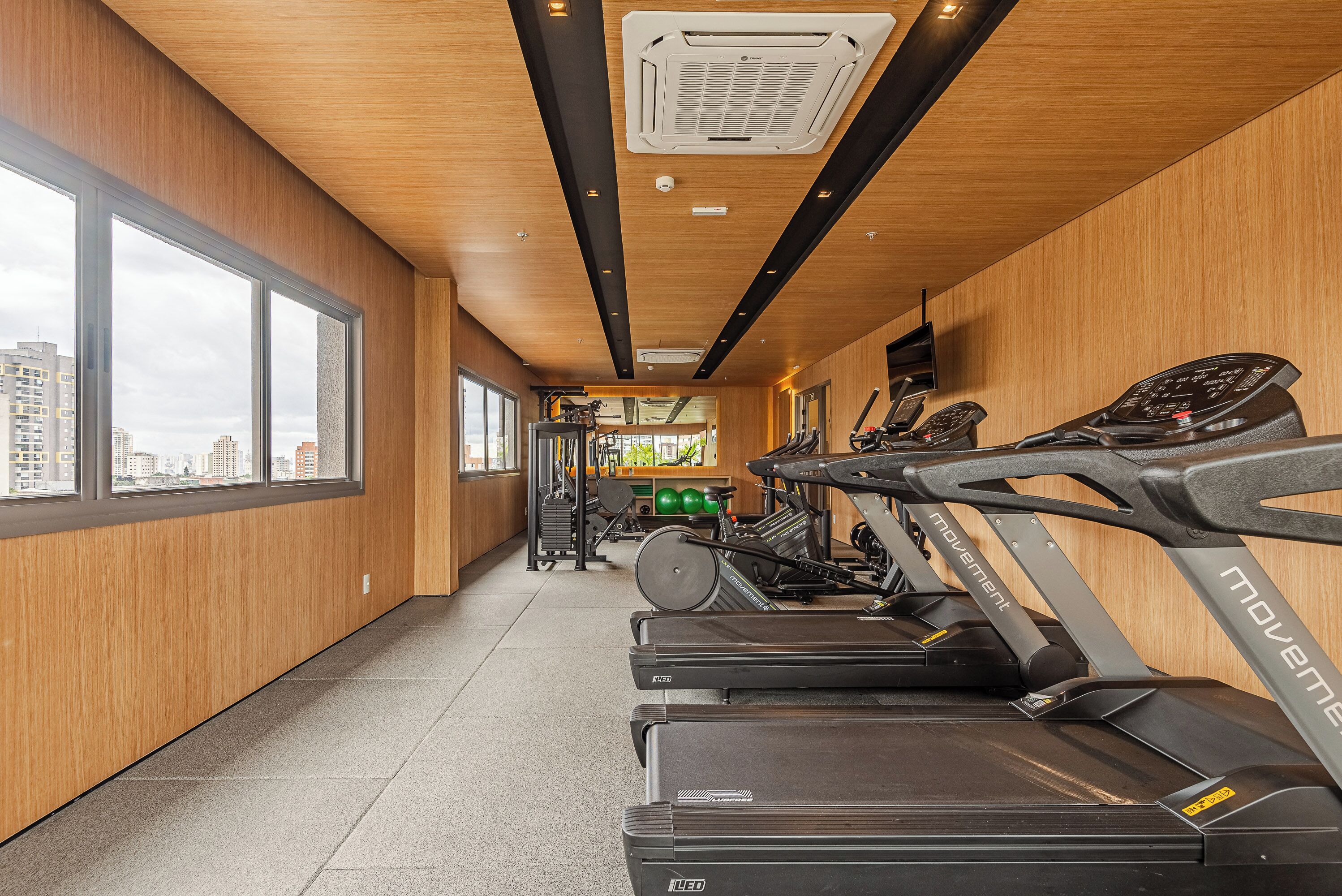 Une salle de sport équipée de tapis de course, d’appareils de musculation et offrant une vue sur la ville grâce à de grandes fenêtres.
