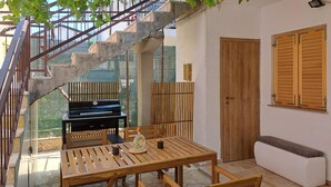 A patio with a wooden table and chairs, a barbecue grill, a wooden bench, and a staircase leading to an upper level.