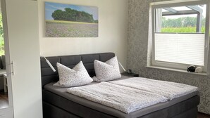 Une chambre avec un grand lit, une table de chevet, une fenêtre avec des stores, une porte et un tableau au mur.