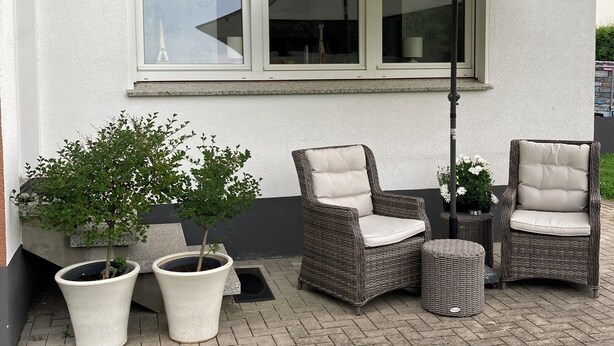 Un espace extérieur aménagé avec des chaises en osier et un parasol blanc, des plantes en pot et un bâtiment doté de grandes fenêtres.