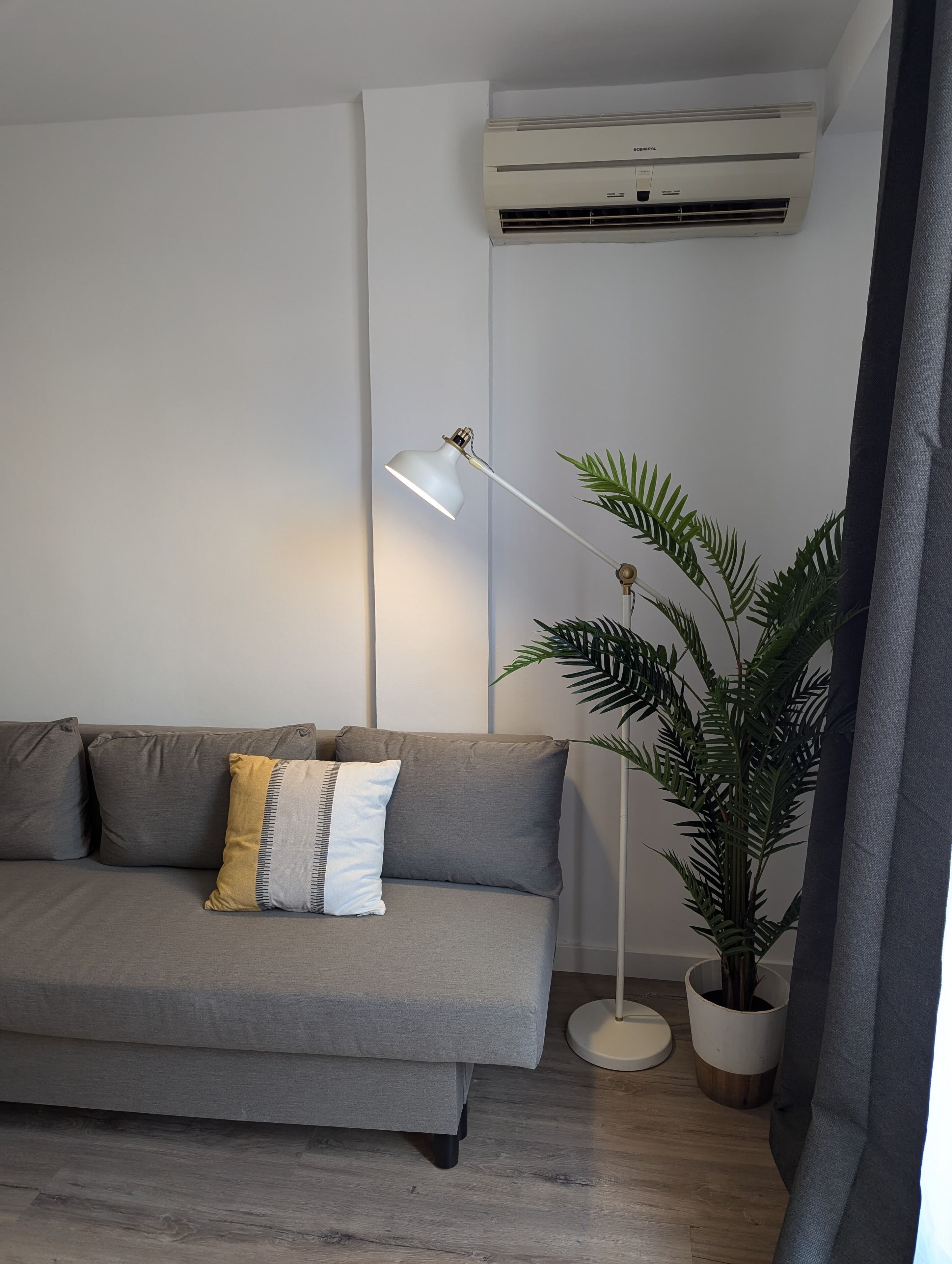 Een moderne woonkamer met een grijze bank, een witte vloerlamp, een potplant en een airconditioning op de muur.