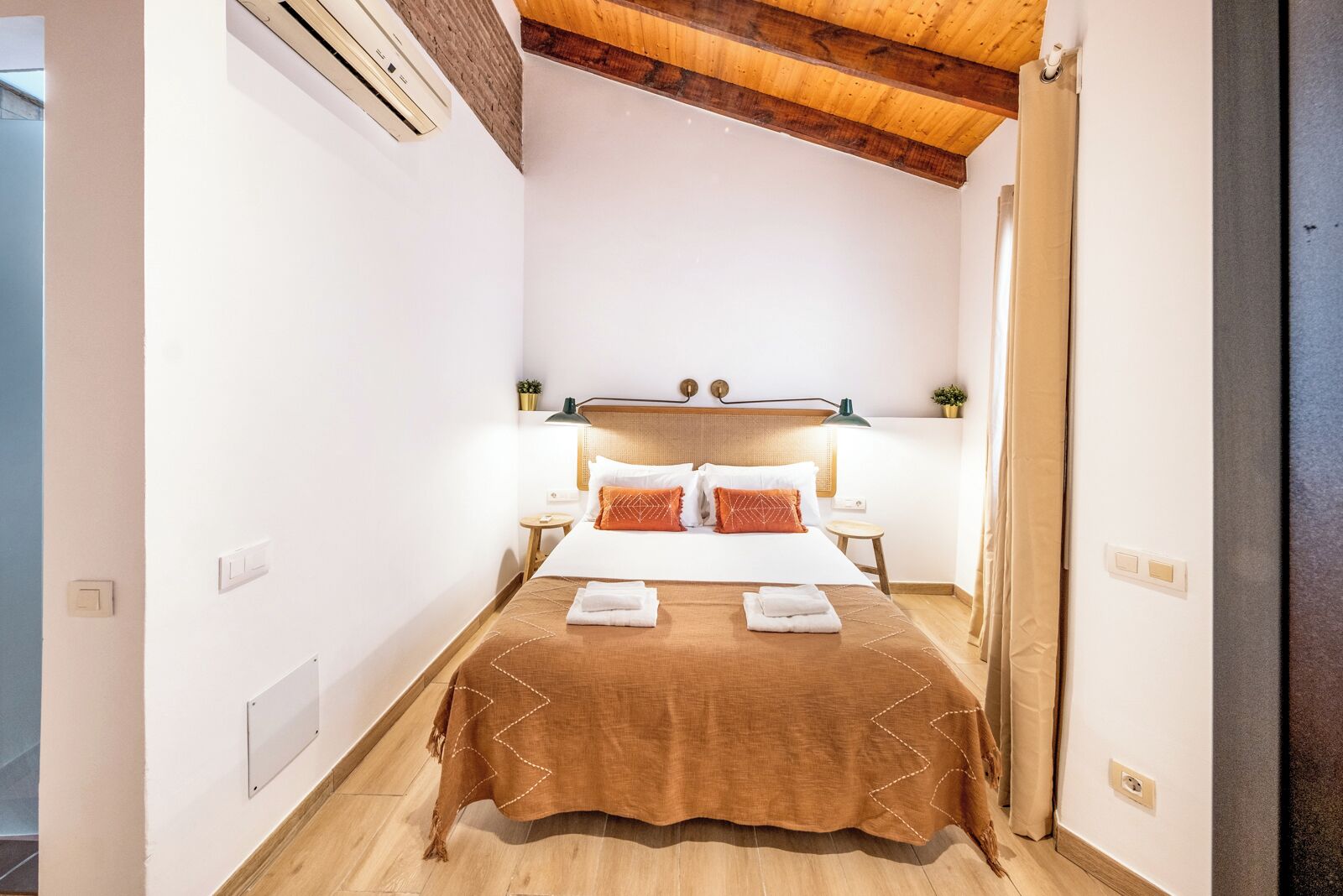 A narrow hotel room with a wooden ceiling, a bed with a brown bedspread, two bedside tables with lamps, and a small potted plant on the nightstand.