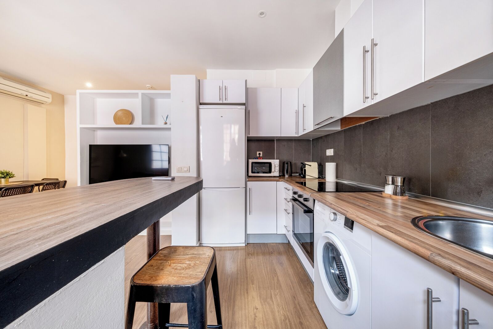 A modern kitchen with white cabinets, a wooden countertop, a built-in washing machine, and a dining area with a table and chairs.