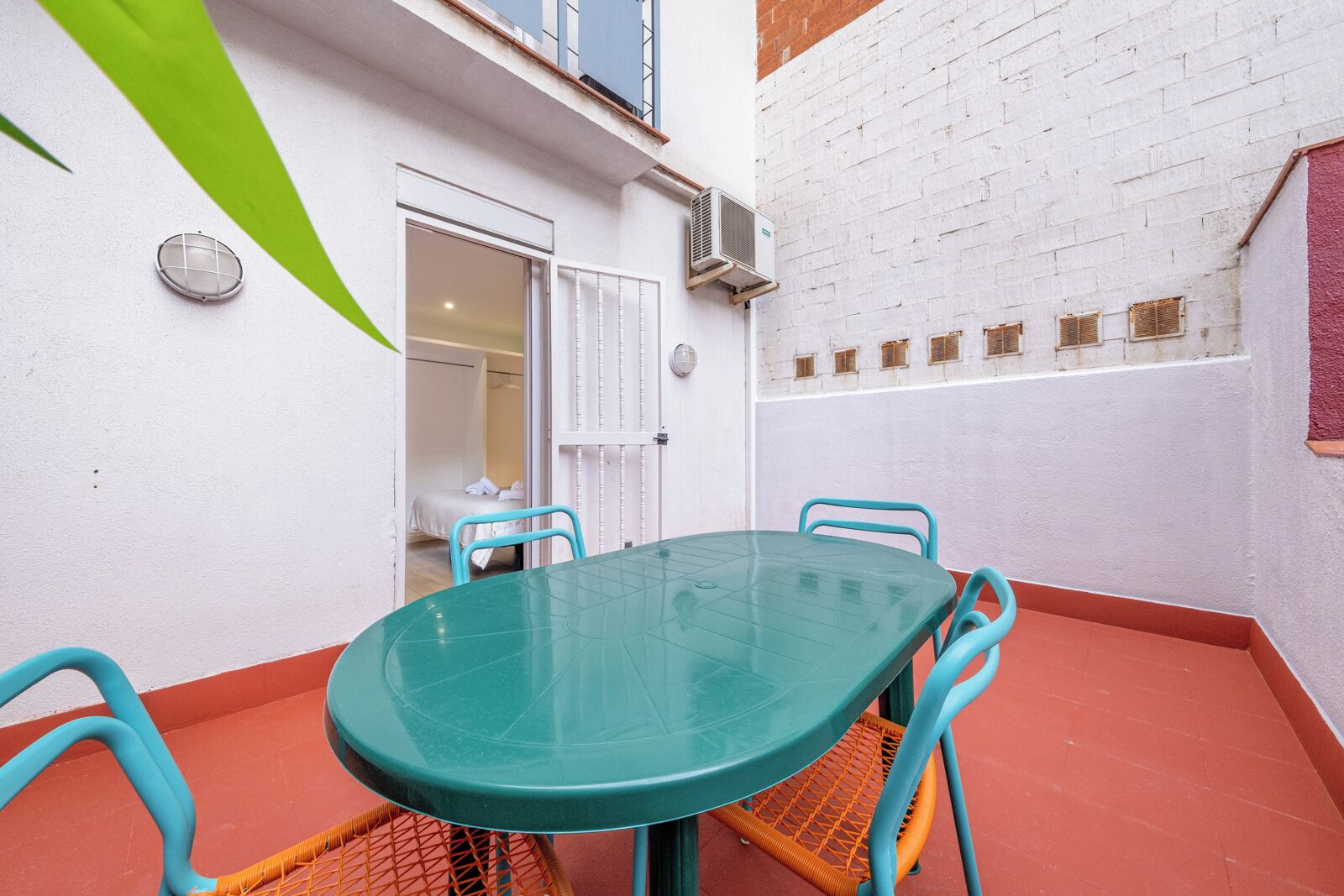 A small outdoor patio with a green table and four chairs, a door leading to a room, and a white brick wall with a window.