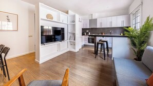 Une cuisine moderne avec des meubles blancs, un four encastré et un grand téléviseur. Le salon est équipé de chaises en bois et d’un canapé gris.