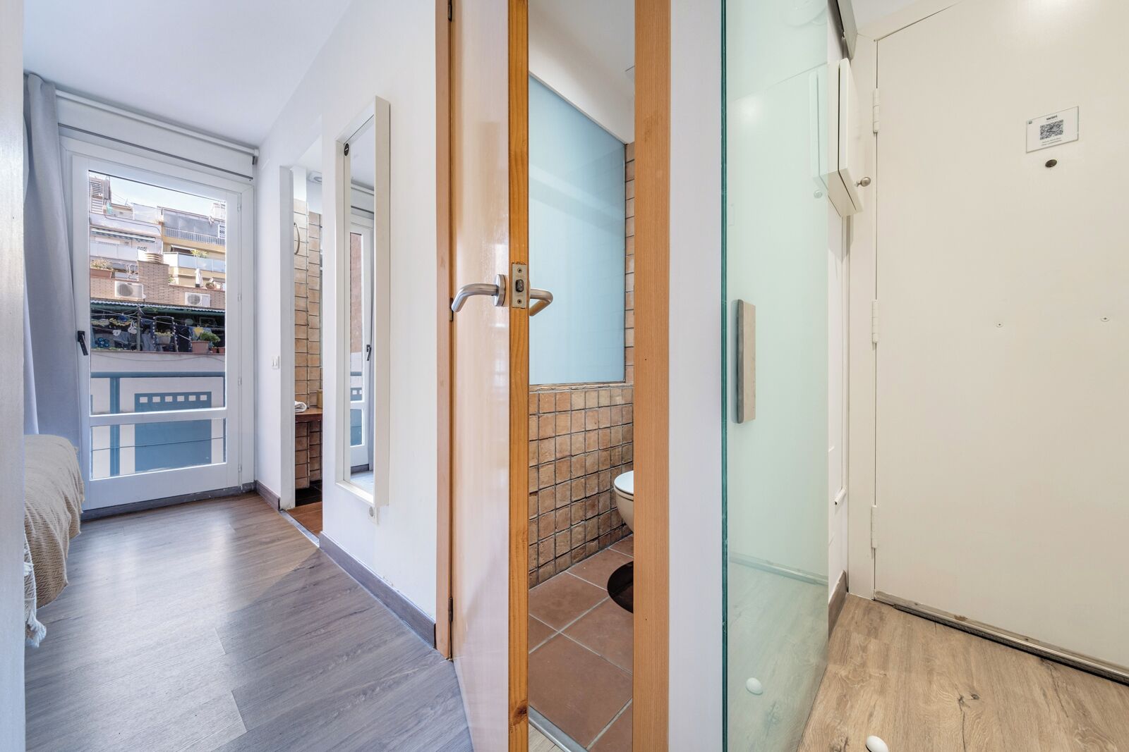 Un pasillo con una puerta corredera de vidrio que da a un balcón, una puerta de madera y una puerta de vidrio hacia el baño.