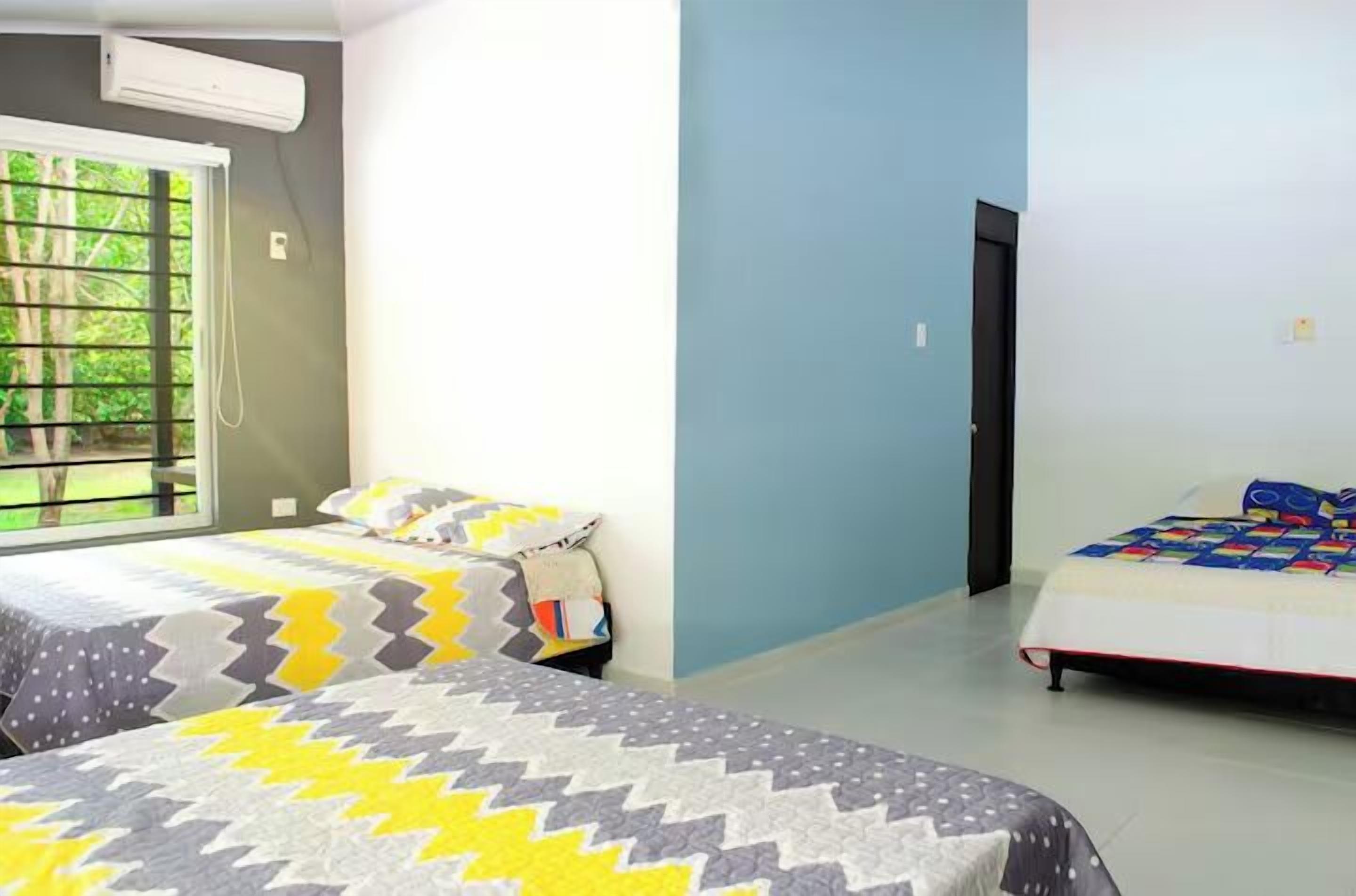 A bedroom with two beds, one with a patterned bedspread and the other with a solid color bedspread. There is a window with a view of greenery outside, and a door leading to another room.