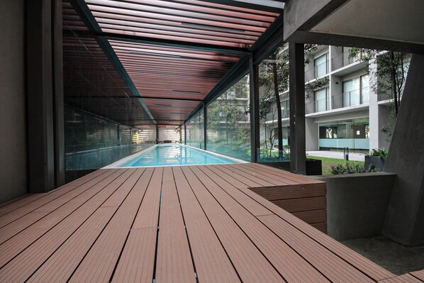 A modern indoor swimming pool with a wooden deck, large windows, and a view of a building.