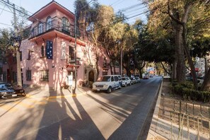 A pink building with a balcony, parked cars, and trees along the street.