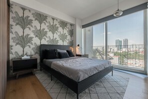 A modern bedroom with a large bed, a nightstand, a TV, and a city view through large windows.