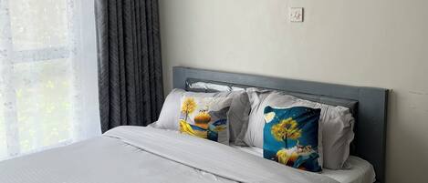 A hotel room with a bed, pillows, a bedside table, a window with curtains, and two framed pictures on the wall.