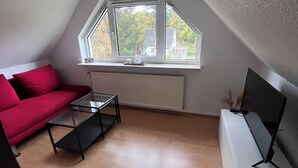 Et loftstudie med skrå vægge, en rød sofa, et glasbord og et fjernsyn på et hvidt skab.