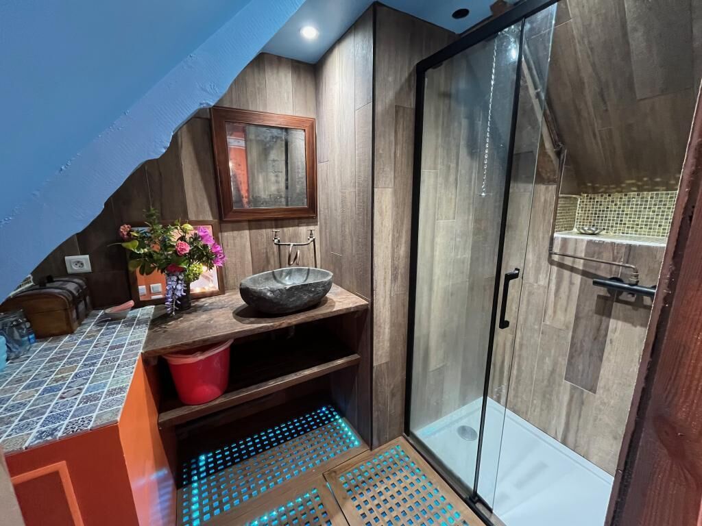 A bathroom with a shower, a sink with a bowl, a mirror, and a wooden shelf.