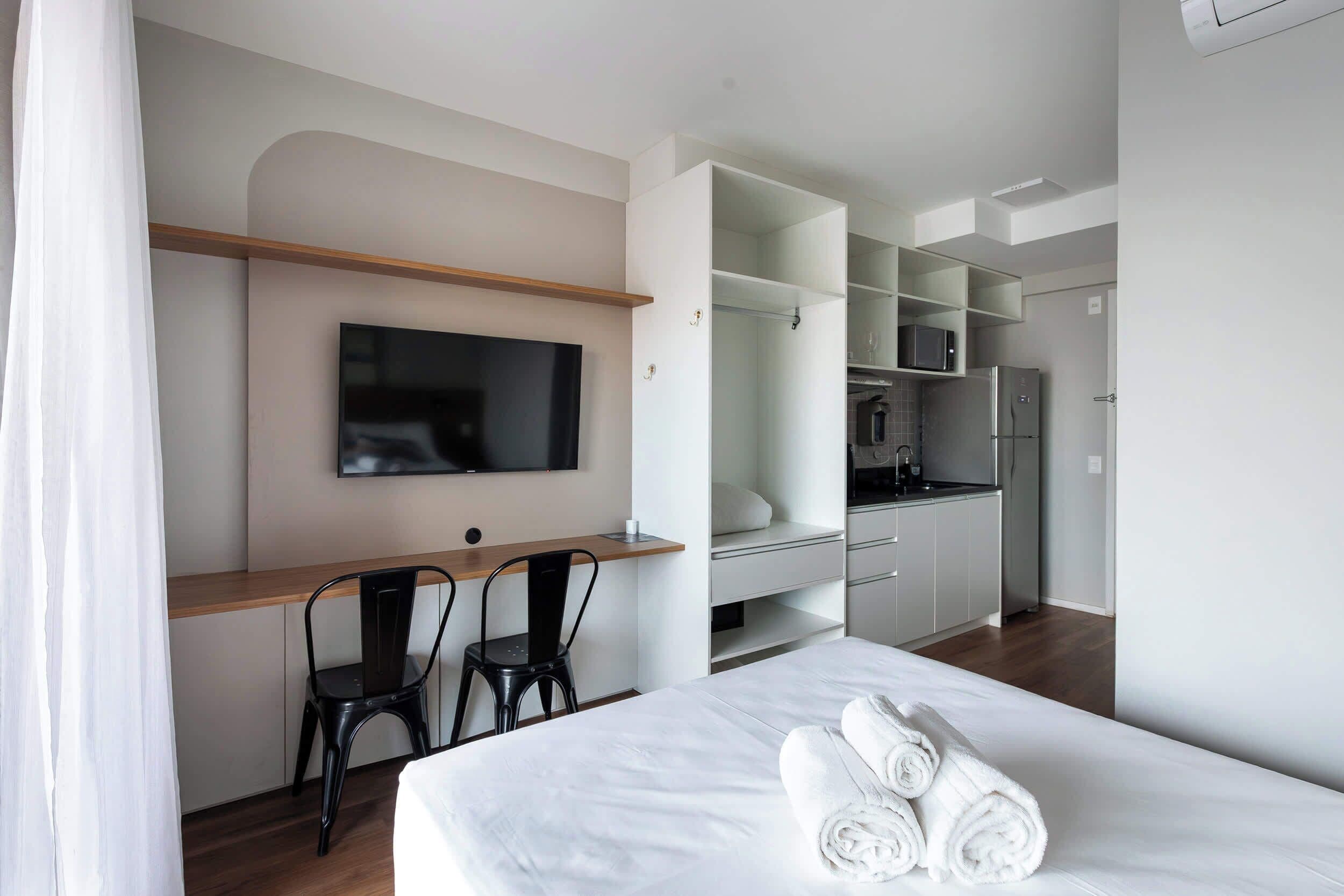 A hotel room with a bed, a desk, two chairs, a television, a kitchenette, and a bathroom.