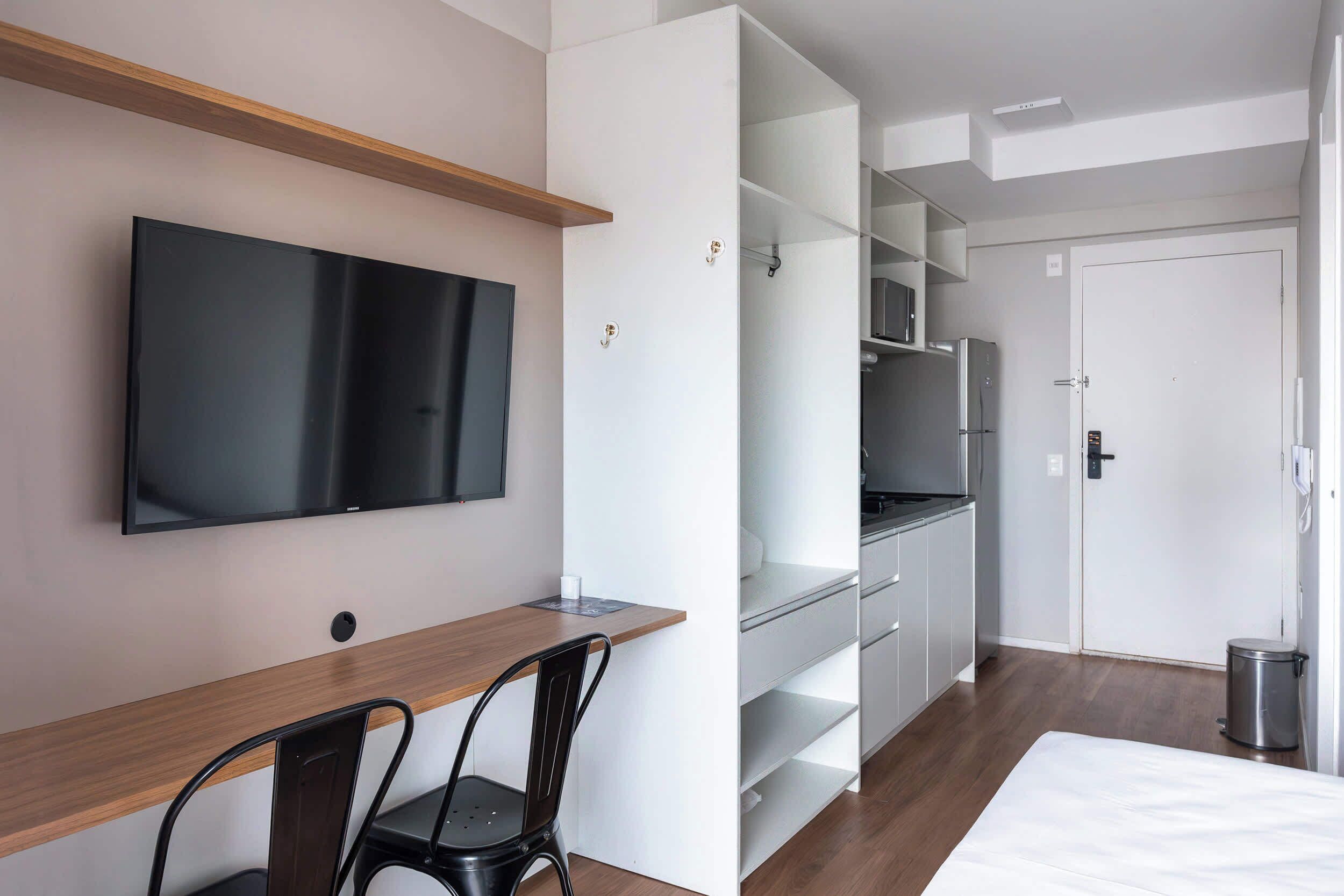 A modern hotel room with a flat-screen TV, a wooden desk, a chair, a bed, a wardrobe, and a trash bin.