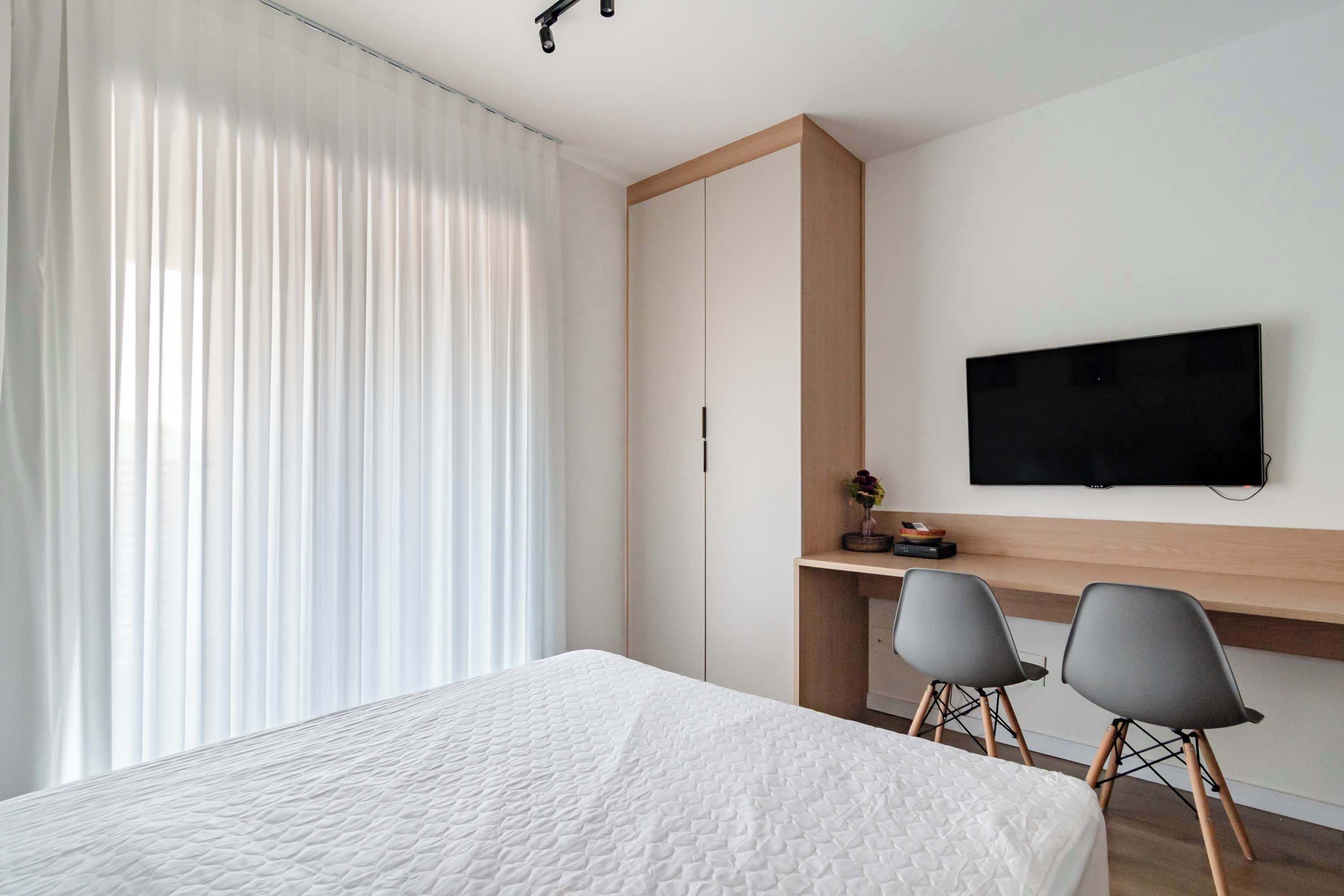 A hotel room with a bed, a TV mounted on the wall, a desk with two chairs, and a large window with curtains.