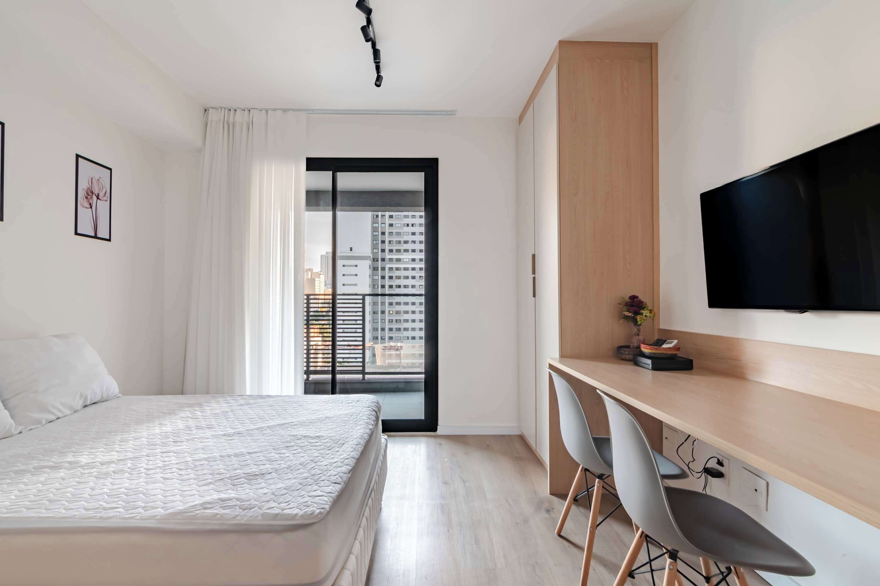 A modern hotel room with a bed, a desk with chairs, a flat-screen TV, and a view of a cityscape through a sliding door.