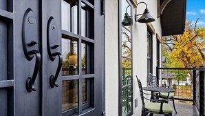 Une porte noire avec une fenêtre, un balcon avec une table et des chaises, et une vue sur les arbres.